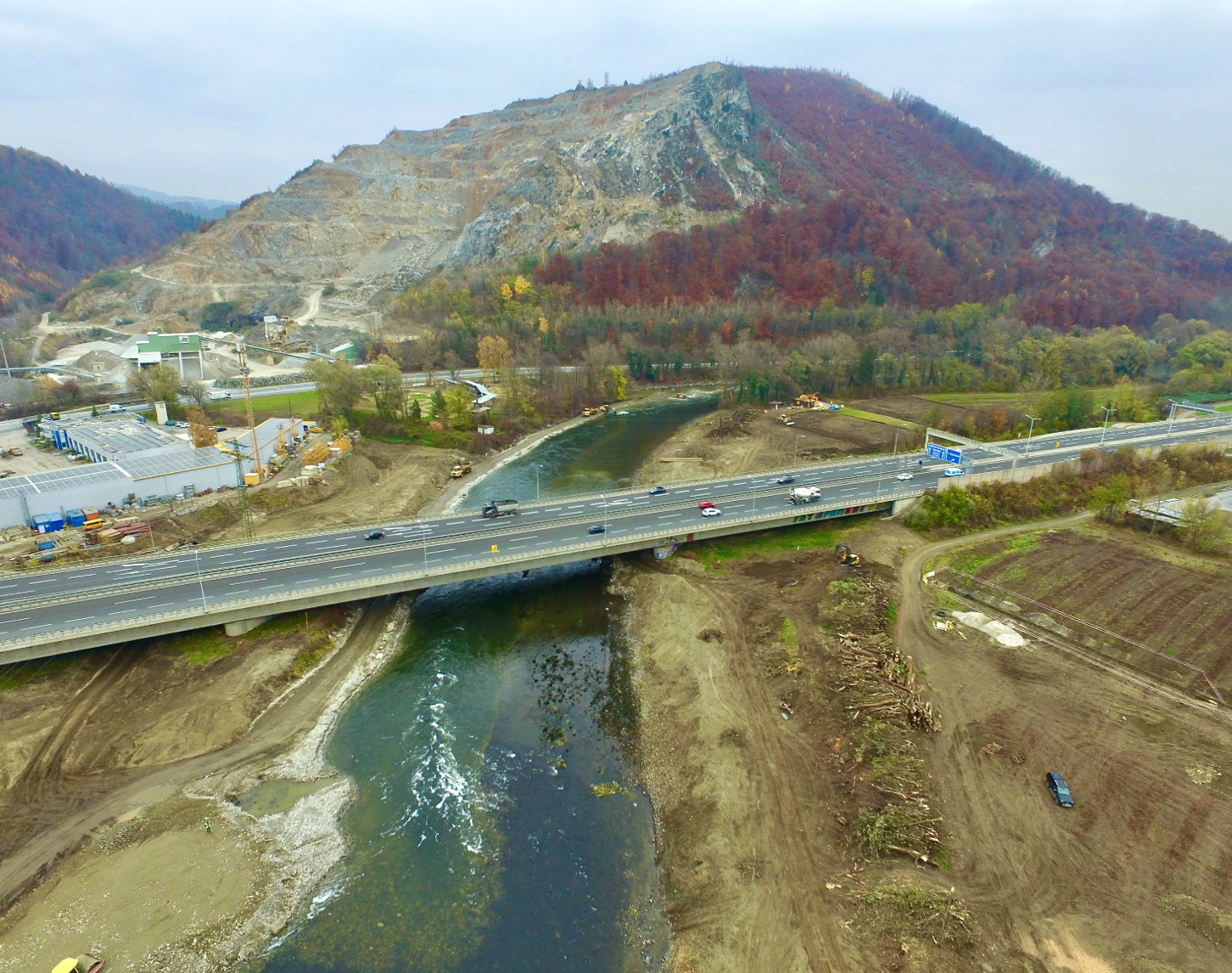 Luftaufnahme der Frühphase der Baustelle am Murkraftwerk Gratkorn. Das Bild zeigt die Bauvorbereitung mit einer Autobahnbrücke über den Fluss Mur, umgeben von entwaldeten Hängen und Anfangsarbeiten auf dem weitläufigen Gelände.
