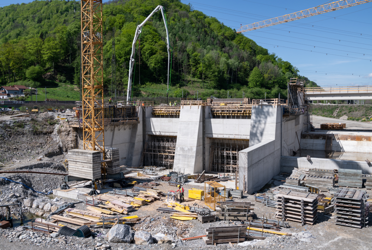 Header-Bild des Baufortschritts vom Mai 2023 am Murkraftwerk Gratkorn, zeigt massive Betonstrukturen, die im Bau sind, mit einem großen gelben Kran, Betonpumpen und verstreuten Baumaterialien.