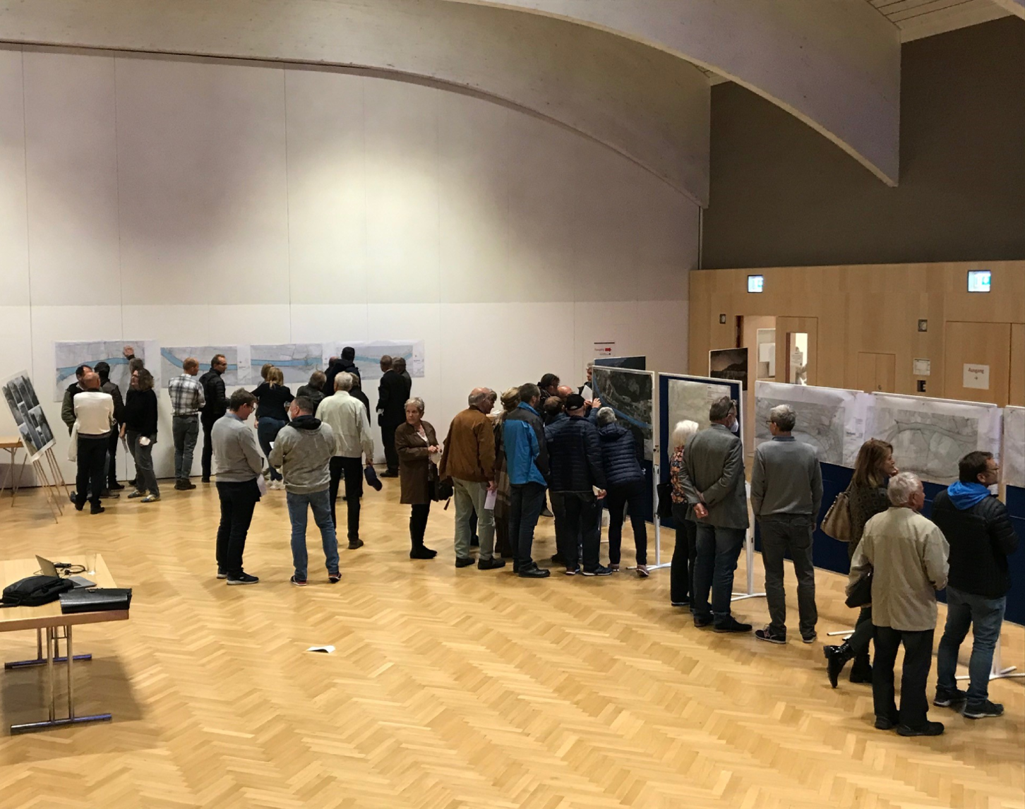 Bild von einem Informationstag zum Murkraftwerksprojekt Gratkorn mit Anrainerinnen und Anrainern, die sich in einem Raum mit Holzboden versammelt haben, um Ausstellungen und Informationsplakate zu betrachten.
