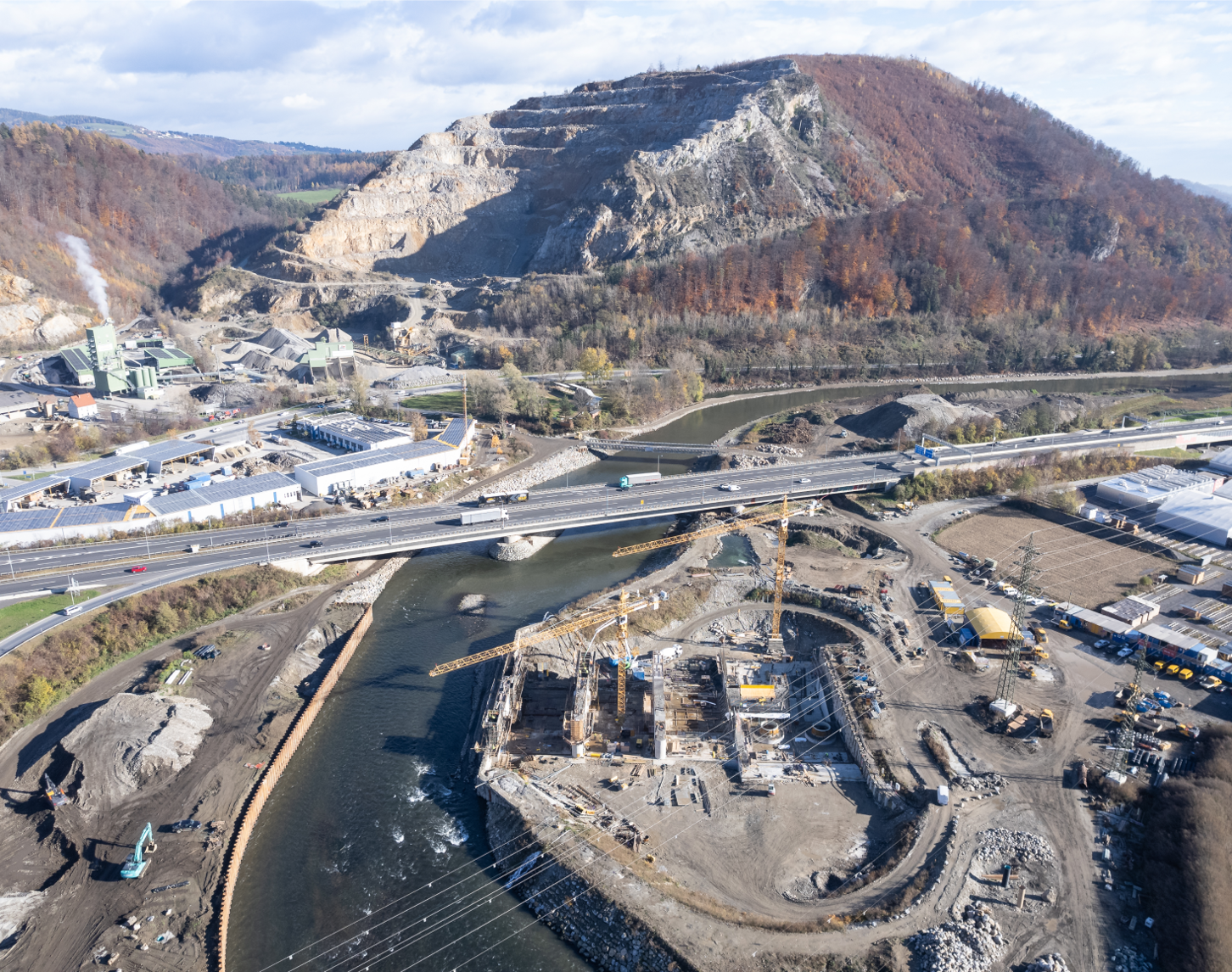 Luftaufnahme des Baufortschritts am Murkraftwerk Gratkorn im Jahr 2022 zeigt die Baustelle mit Kränen und halbfertigen Betonstrukturen nahe der Autobahn und dem Flusslauf der Mur, mit Steinbruchhügeln im Hintergrund.