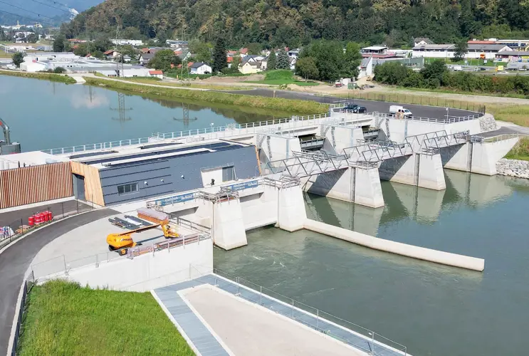 Luftaufnahme des neu errichteten Murkraftwerks Kraftwerk Gratkorn. Man sieht den Wasserdurchlass, das Betriebsgebäude und das Wehr mit geöffneten Schleusentoren in einer grünen Landschaft mit einem Fluss.