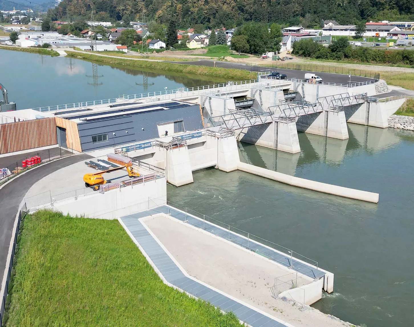 Luftaufnahme des neu errichteten Murkraftwerks Kraftwerk Gratkorn. Man sieht den Wasserdurchlass, das Betriebsgebäude und das Wehr mit geöffneten Schleusentoren in einer grünen Landschaft mit einem Fluss.