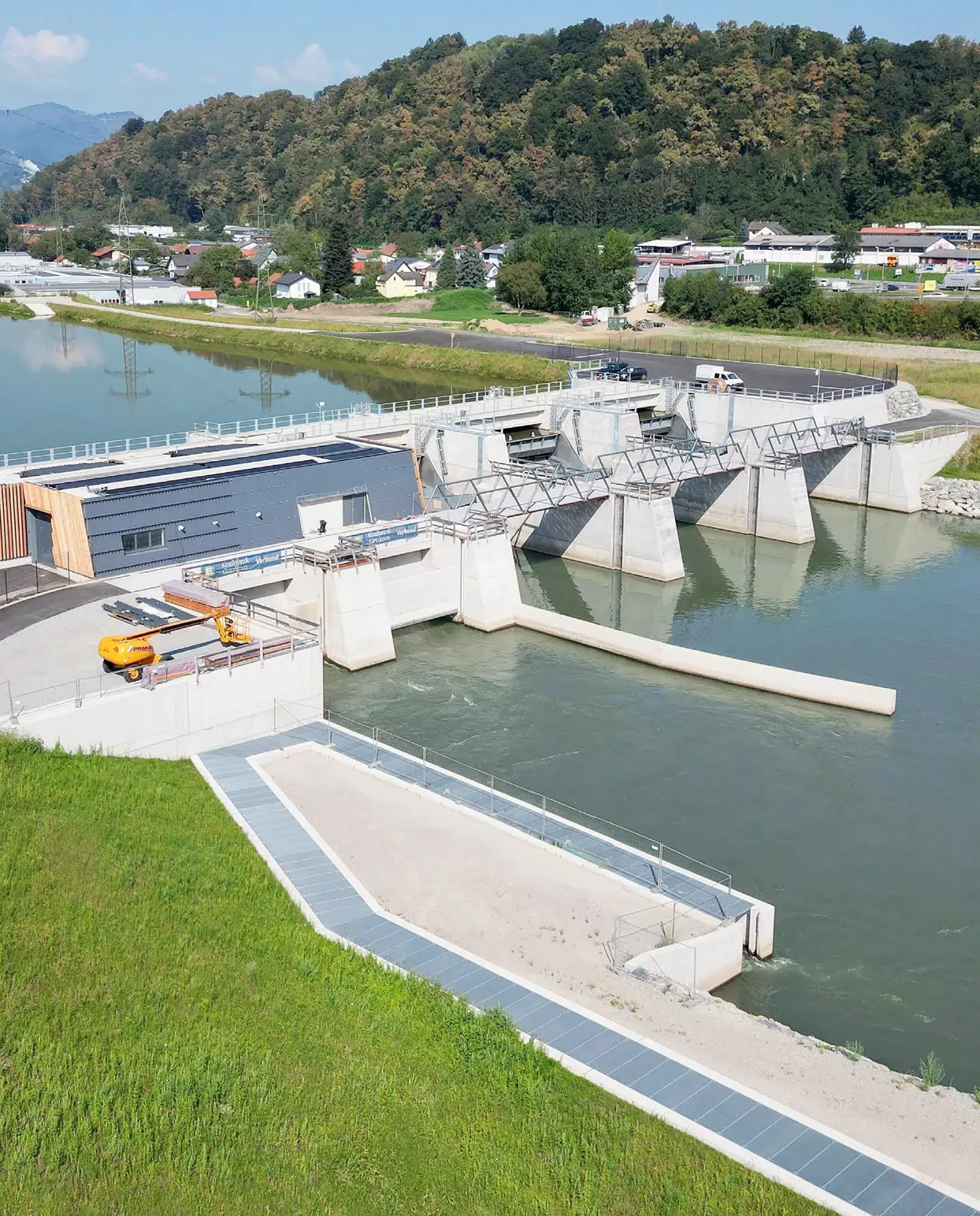 Luftaufnahme des neu errichteten Murkraftwerks Kraftwerk Gratkorn. Man sieht den Wasserdurchlass, das Betriebsgebäude und das Wehr mit geöffneten Schleusentoren in einer grünen Landschaft mit einem Fluss.